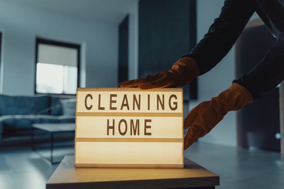 A person wearing brown rubber cleaning gloves is adjusting a small illuminated sign on a wooden surface that reads 'CLEANING HOME'. The background features a modern living room with a sofa, a low coff