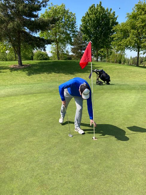 A person on a golf course bending down to align a putt near the green, wearing a blue jacket, white pants, and a white cap. The green surface shows well-maintained, short grass, and a red flagstick is